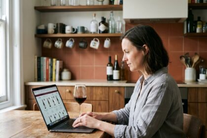 Femme achetant du vin en ligne sur son ordinateur portable dans une cuisine moderne avec un verre de vin rouge posé à côté
